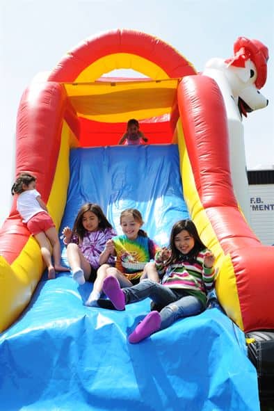 An inflatable slide with four girls playing on it. The slide is primarily blue and yellow, with a red arch over the top. One girl is at the top of the slide, while the other three are sitting at the bottom, smiling.